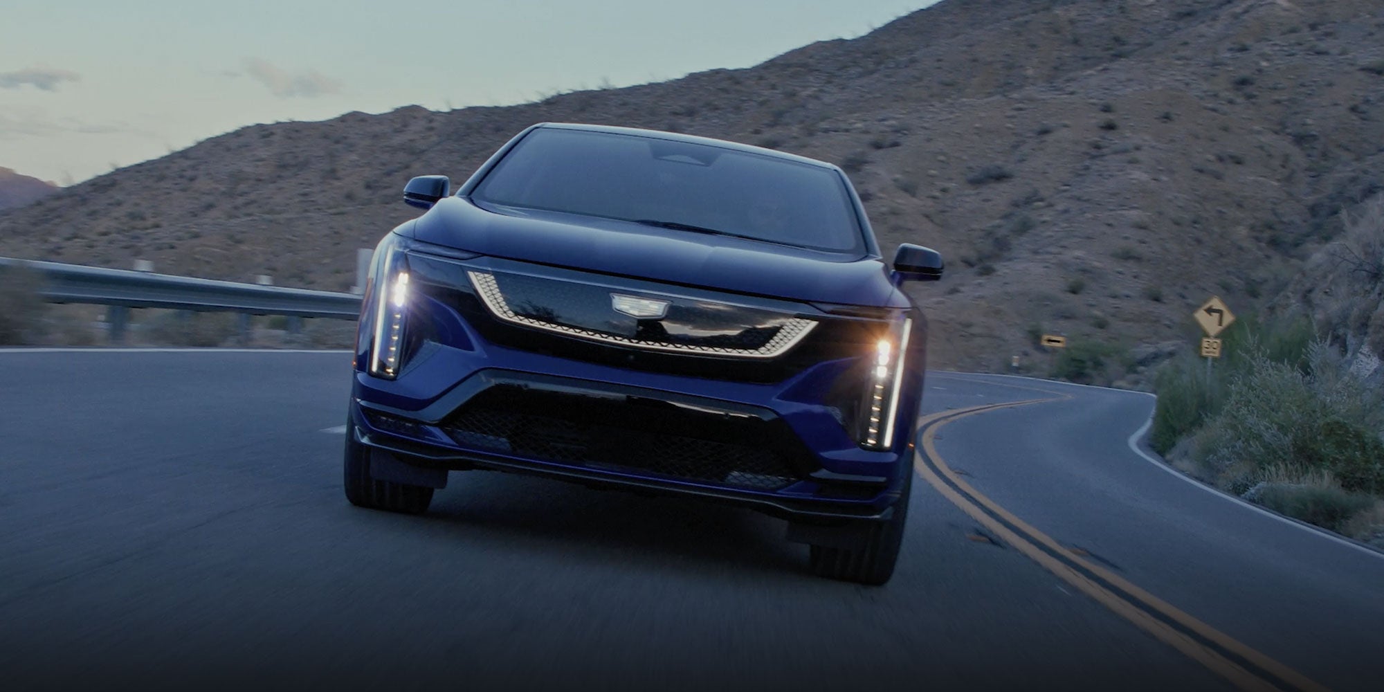 Head On View of a Blue Cadillac OPTIQ-V Driving on a Mountain Road with the Headlights On